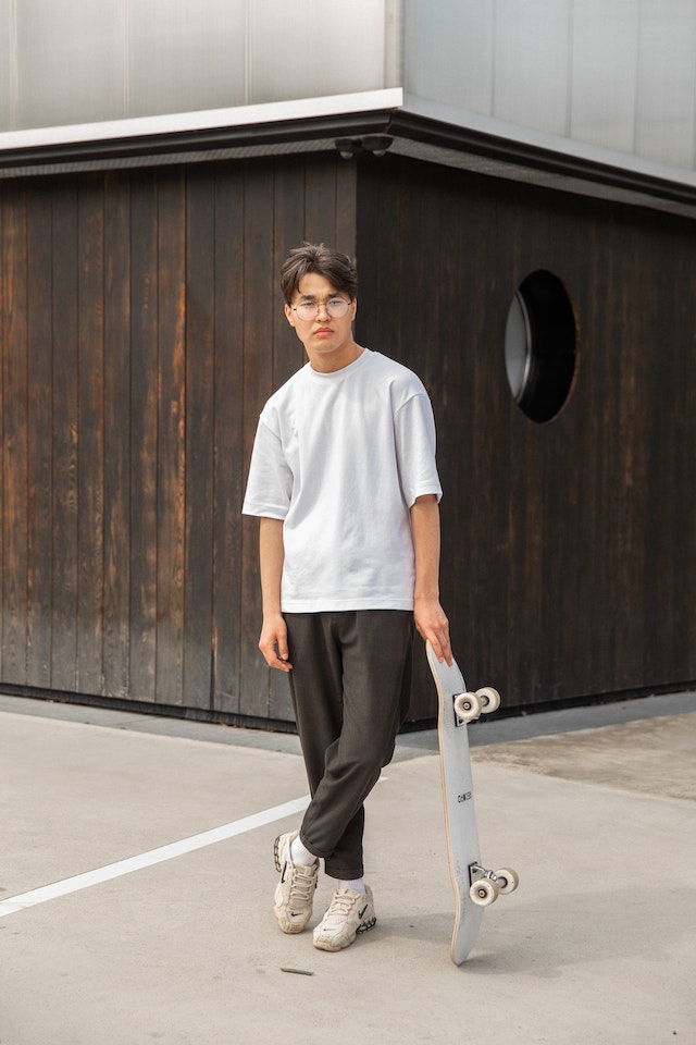 K-Fashion: Mann mit weißem T-Shirt und schwarzer Jogginghose lehnt sich an Skateboard an